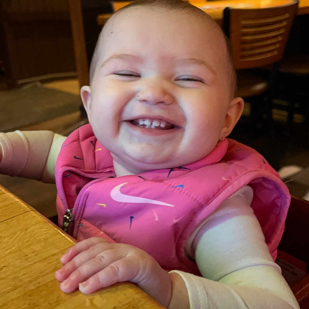 Melanie is registered to the contest to win money with this photo: baby, cheerful, child, clothing, cute, face, happy, high_chair, indoor, infant, joyful, person, pink_vest, portrait, restaurant, seated, sleeves, smiling, table, wood