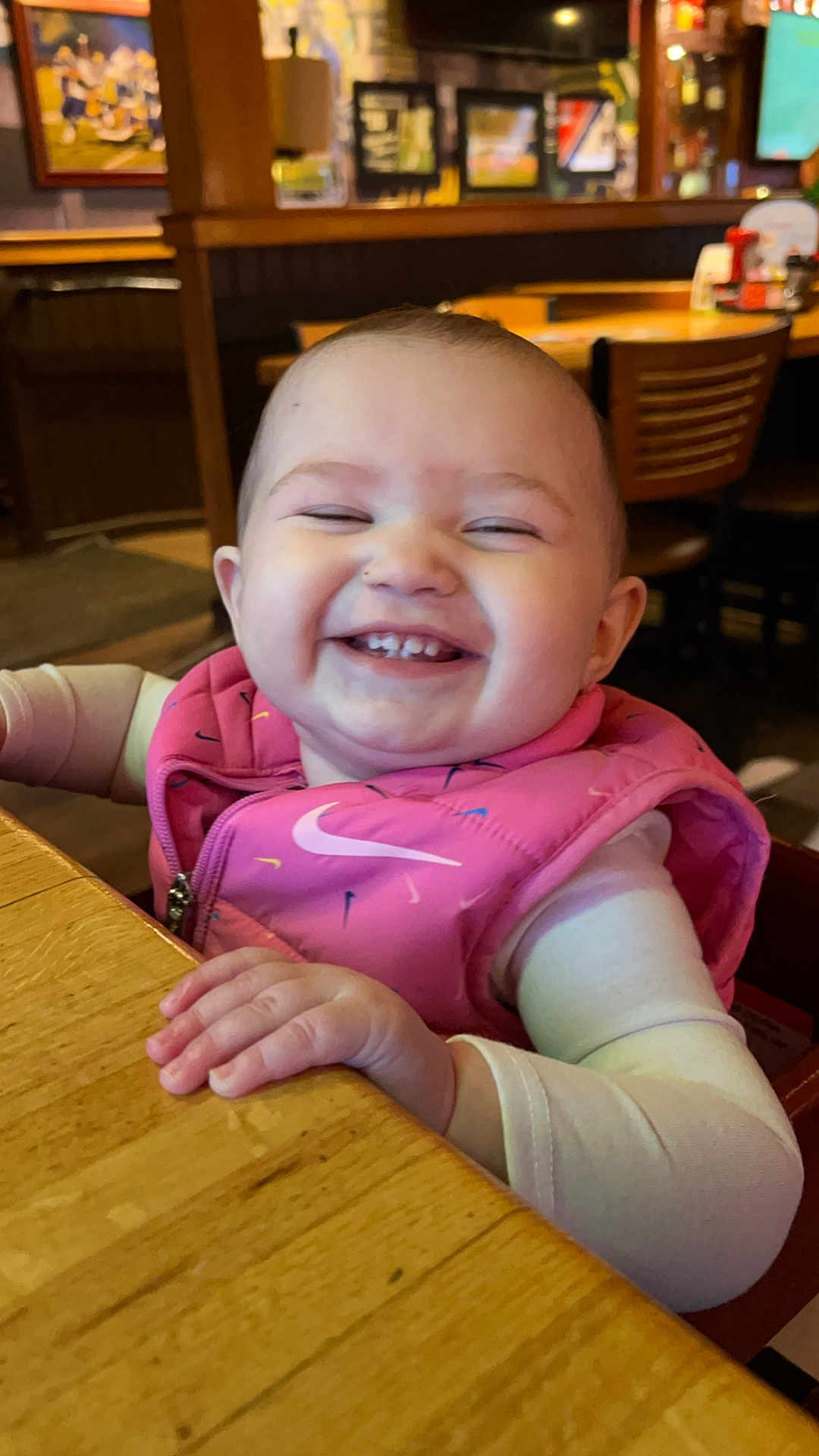 Melanie is registered to the contest to win money with this photo: baby, child, smiling, happy, pink_vest, table, wood, indoor, restaurant, high_chair, person, face, clothing, sleeves, cute, joyful, cheerful, infant, seated, portrait
