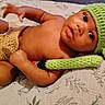 baby, child, infant, portrait, lying_down, bed, blanket, knitted_hat, green_hat, bunny_ears, clothing, diaper_cover, brown_skin, wide_eyes, face, eyes, lips, indoors, patterned_bedding, cute