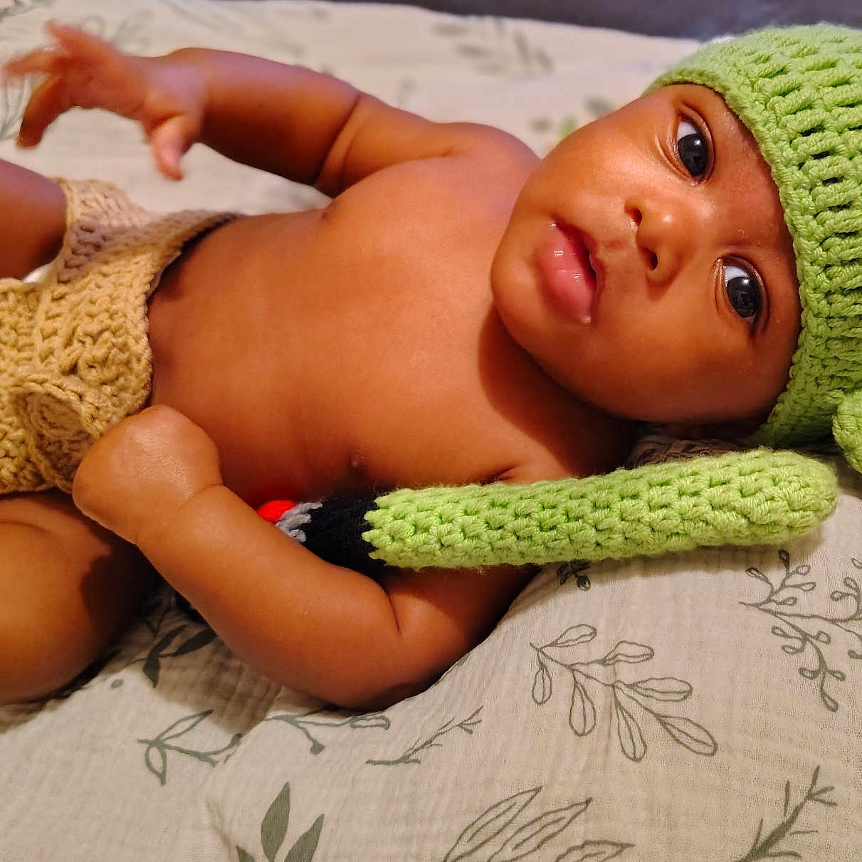 Zamar is registered to the contest to win money with this photo: baby, child, infant, portrait, lying_down, bed, blanket, knitted_hat, green_hat, bunny_ears, clothing, diaper_cover, brown_skin, wide_eyes, face, eyes, lips, indoors, patterned_bedding, cute