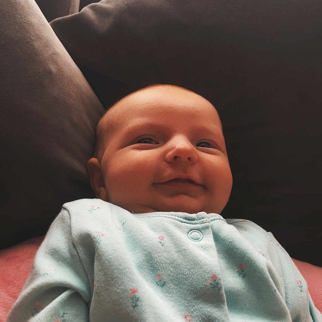 Nova-Mae is registered to the contest to win money with this photo: baby, child, clothing, comfortable, cozy, cushion, cute, face, floral_print, happy, head, home, indoor, infant, onesie, portrait, relaxed, smiling, sofa, soft_lighting