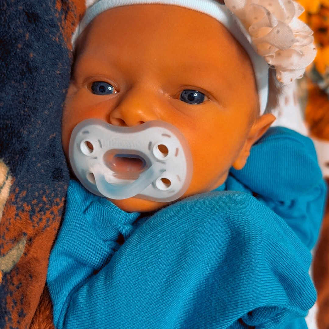 Mylee is registered to the contest to win money with this photo: baby, blanket, blue_outfit, child, close_up, cute, eyes, face, flower, hand, headband, infant, lying_down, newborn, pacifier, patterned_blanket, skin, sleepwear, soft_texture, warm_colors