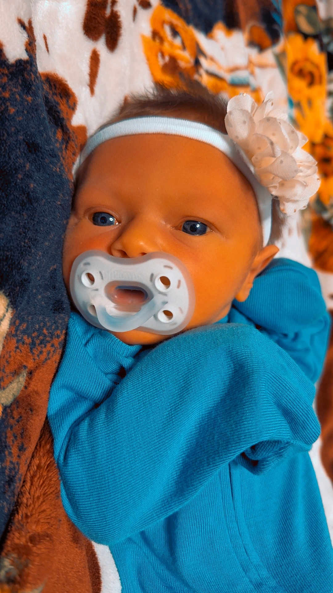 Mylee is registered to the contest to win money with this photo: baby, pacifier, blue_outfit, headband, flower, blanket, close_up, infant, child, face, cute, sleepwear, patterned_blanket, newborn, skin, eyes, hand, lying_down, warm_colors, soft_texture
