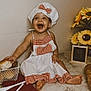 baby, child, chef_hat, apron, smiling, happy, bread, cookie, kitchen_utensils, marble_countertop, sunflowers, flower_arrangement, food, sitting, cute, infant, table, red_checkered_pattern, playful, indoor