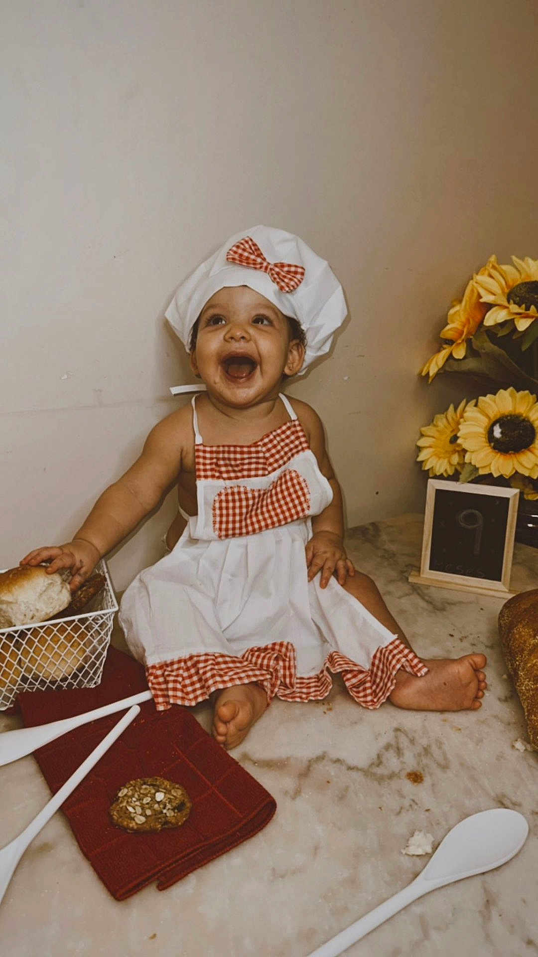 Rebecca is registered to the contest to win money with this photo: baby, child, chef_hat, apron, smiling, happy, bread, cookie, kitchen_utensils, marble_countertop, sunflowers, flower_arrangement, food, sitting, cute, infant, table, red_checkered_pattern, playful, indoor