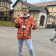 Kenzo participe au concours pour gagner de l'argent avec cette photo : child, face_paint, glasses, orange_jacket, dinosaur_pattern, grey_sweatpants, black_shoes, outdoor, pavement, medieval_buildings, playful_pose, smiling, person, daylight, trees, bench, wooden_wheel, grass, overcast_sky, casual_clothing