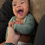 Kyngstun is registered to the contest to win money with this photo: baby, laughing, hand, tattoo, watch, diaper, couch, phone, green_shirt, happy, child, infant, person, indoor, smile, holding, arm, skin, closeup, casual