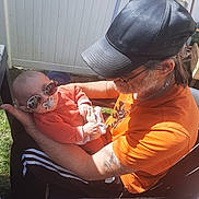 Elio a rejoint le concours — aidez-le/la à gagner de superbes lots ! baby, sunglasses, pacifier, man, beard, cap, orange_shirt, outdoor, sunlight, fence, chair, grass, tattoo, glasses, holding, casual_clothing, relaxing, portrait, person, daylight