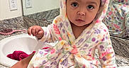 Asiya is registered to the contest to win money with this photo: baby, bathrobe, bathroom, bow, child, clothing, countertop, curious, cute, face, furniture, hood, indoor, mirror, person, portrait, reflection, sink, sitting, towel