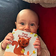 Léo a rejoint le concours — aidez-le/la à gagner de superbes lots ! baby, infant, board_book, book, hands, eyes, face, chewing, couch, blanket, red_cushion, blue_fabric, portrait, close_up, indoor, cozy, cute, small_person, toy, sitting