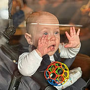 Léo participe au concours pour gagner de l'argent avec cette photo : baby, infant, stroller, car_seat, toy_ball, hands, face, blue_eyes, surprised_expression, window_glass, reflection, blurred_background, soft_light, portrait, cute, sitting, onesie, seat_buckle, playtime, indoor