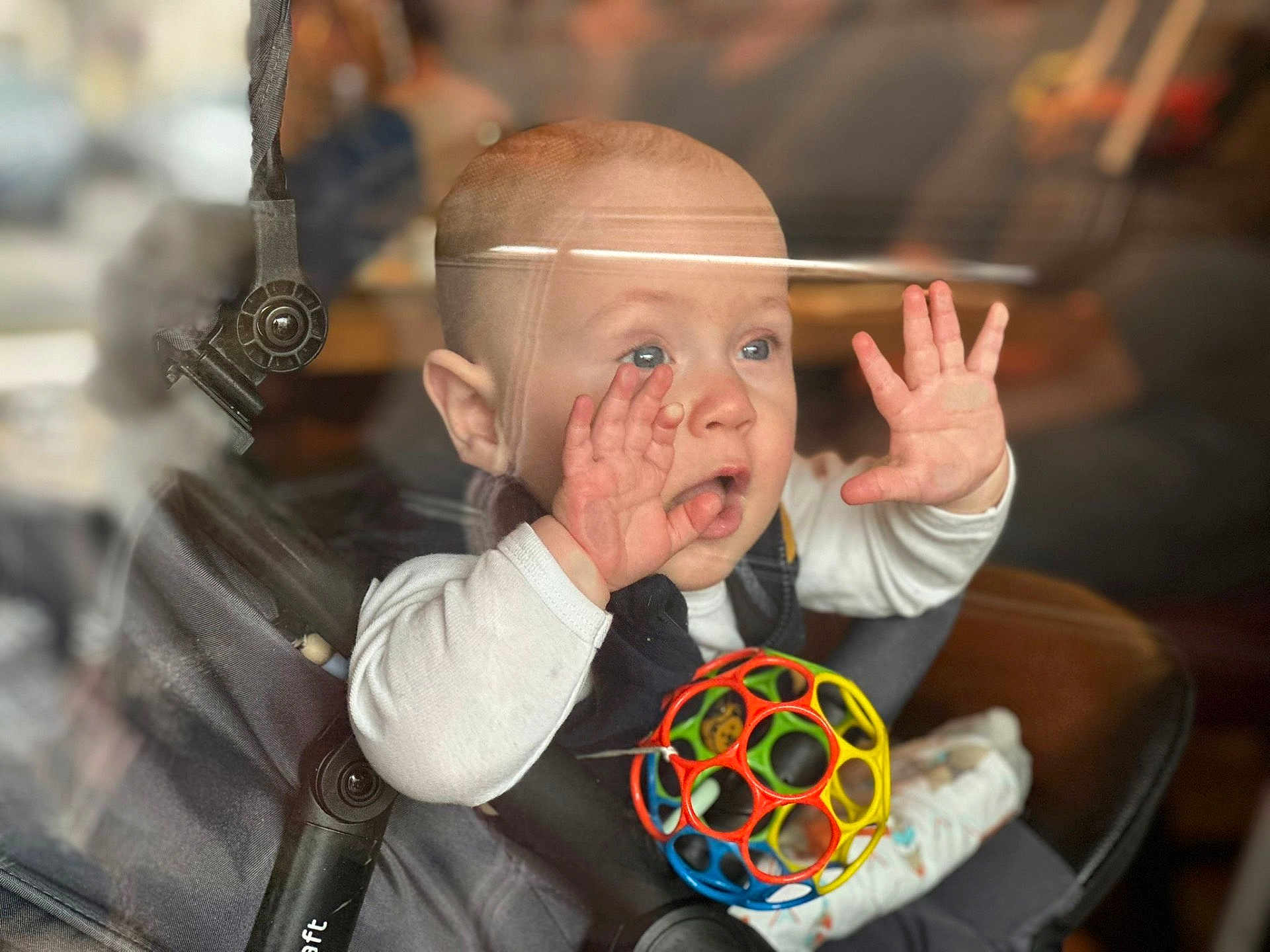 Léo participe au concours pour gagner de l'argent avec cette photo : baby, infant, stroller, car_seat, toy_ball, hands, face, blue_eyes, surprised_expression, window_glass, reflection, blurred_background, soft_light, portrait, cute, sitting, onesie, seat_buckle, playtime, indoor