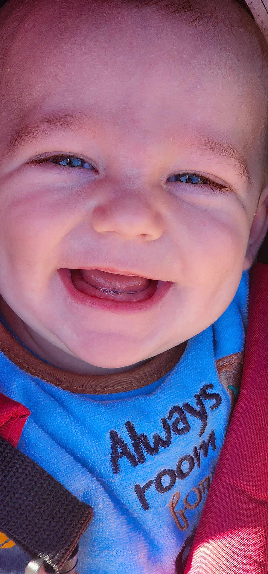 Alister is registered to the contest to win money with this photo: baby, close_up, smile, happy, face, infant, child, blue_clothing, red_strap, teeth, mouth, skin, eyebrows, head, seatbelt, car_seat, outdoor, sunlight, expression, person