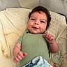 baby, infant, child, smile, quilt, blanket, onesie, green_clothing, curly_hair, lying_down, cute, indoor, soft_texture, bed, happy, face, skin, person, young_child, cozy