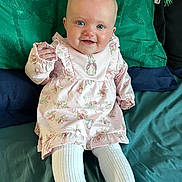 Macie is registered to the contest to win money with this photo: baby, child, smiling, blue_eyes, pink_dress, floral_pattern, tights, white_tights, sitting, bed, pillows, green_pillow, blue_pillow, cute, indoors, portrait, happy, infant, young_child, person