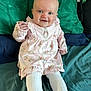 baby, child, smiling, blue_eyes, pink_dress, floral_pattern, tights, white_tights, sitting, bed, pillows, green_pillow, blue_pillow, cute, indoors, portrait, happy, infant, young_child, person