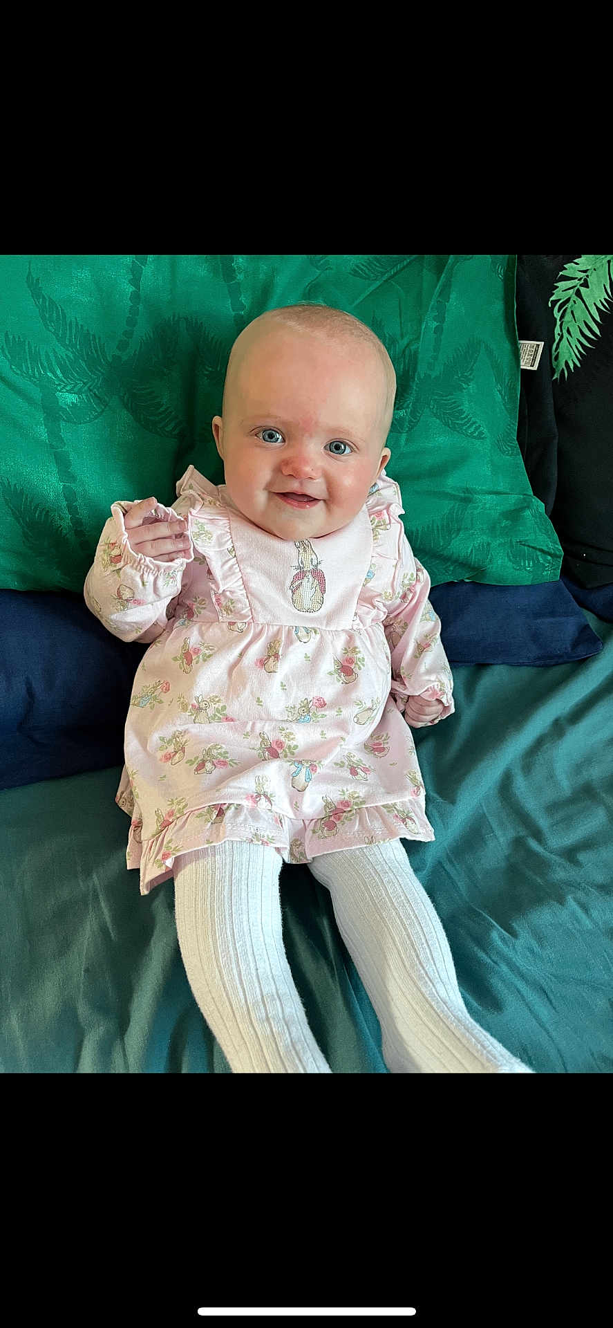 Macie is registered to the contest to win money with this photo: baby, child, smiling, blue_eyes, pink_dress, floral_pattern, tights, white_tights, sitting, bed, pillows, green_pillow, blue_pillow, cute, indoors, portrait, happy, infant, young_child, person