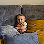 baby, child, pillow, couch, soft, orange_ball, curious, indoor, seated, cozy, plush, furniture, home, cute, infant, comfort, person, casual, relaxed, young