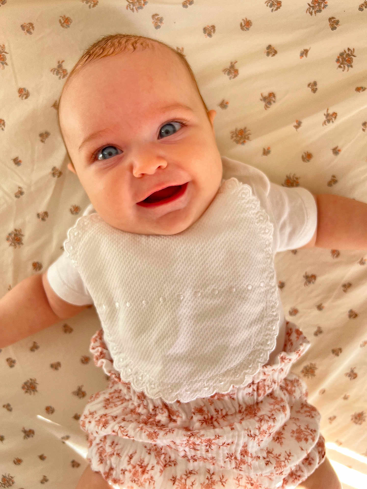 Mily-Rose participe au concours pour gagner de l'argent avec cette photo : baby, smiling, blue_eyes, white_bib, floral_bloomers, lying_down, floral_sheet, cute, happy, infant, portrait, child, clothing, skin, head, face, arms, baby_girl, indoors, soft_light