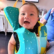 John joined the competition — help win amazing prizes! baby, child, smiling, life_jacket, indoor, person, blue_shirt, happiness, portrait, cute, infant, clothing, colorful, face, young_child, happy, closeup, arm, zipper, background