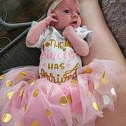 Evie is registered to the contest to win money with this photo: baby, newborn, infant, headband, bow, tutu, pink_tulle, gold_polka_dots, onesie, text_on_clothing, hands, face, eyes, mouth, legs, lap, sofa, blanket, cute, portrait