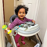 Ariella joined the competition — help win amazing prizes! toddler, child, baby_walker, toy, indoor, smiling, curly_hair, purple_clothing, pink_socks, floor_tile, door, door_handle, baby, playful, person, walker, happy, standing, young_child, home