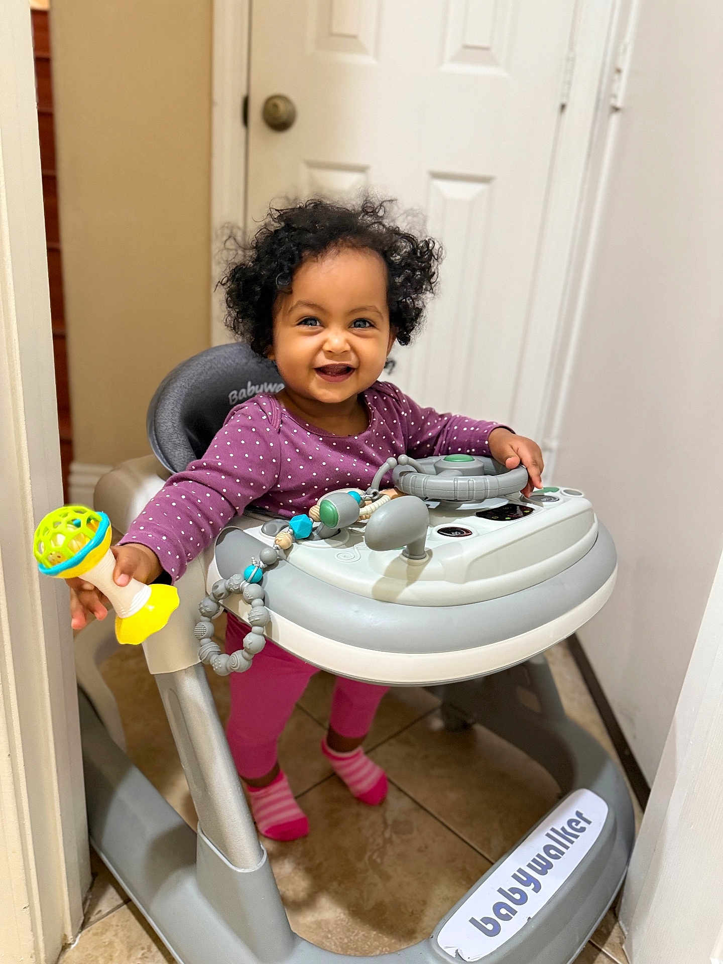 Ariella joined the competition — help win amazing prizes! toddler, child, baby_walker, toy, indoor, smiling, curly_hair, purple_clothing, pink_socks, floor_tile, door, door_handle, baby, playful, person, walker, happy, standing, young_child, home