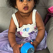 Ariella is registered to the contest to win money with this photo: toddler, child, curly_hair, sippy_cup, purple_skirt, crocheted_top, couch, leather, seated, wide_eyes, expression, indoor, hands, clothing, baby, cute, portrait, skin, furniture, young_child