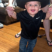 Carter is registered to the contest to win money with this photo: toddler, child, cowboy_hat, black_shirt, jeans, smiling, indoor, wooden_floor, person, holding_hands, adult, living_room, footwear, nike, happy, cute, casual_clothing, home, young_child, playful