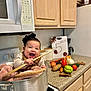 apple, appliance, baby, bow, cabinet, carrot, celebration, child, cooking_pot, countertop, green_pepper, happy, indoor, kitchen, potato, smiling, stove, tomato, vegetables, wooden_spoon
