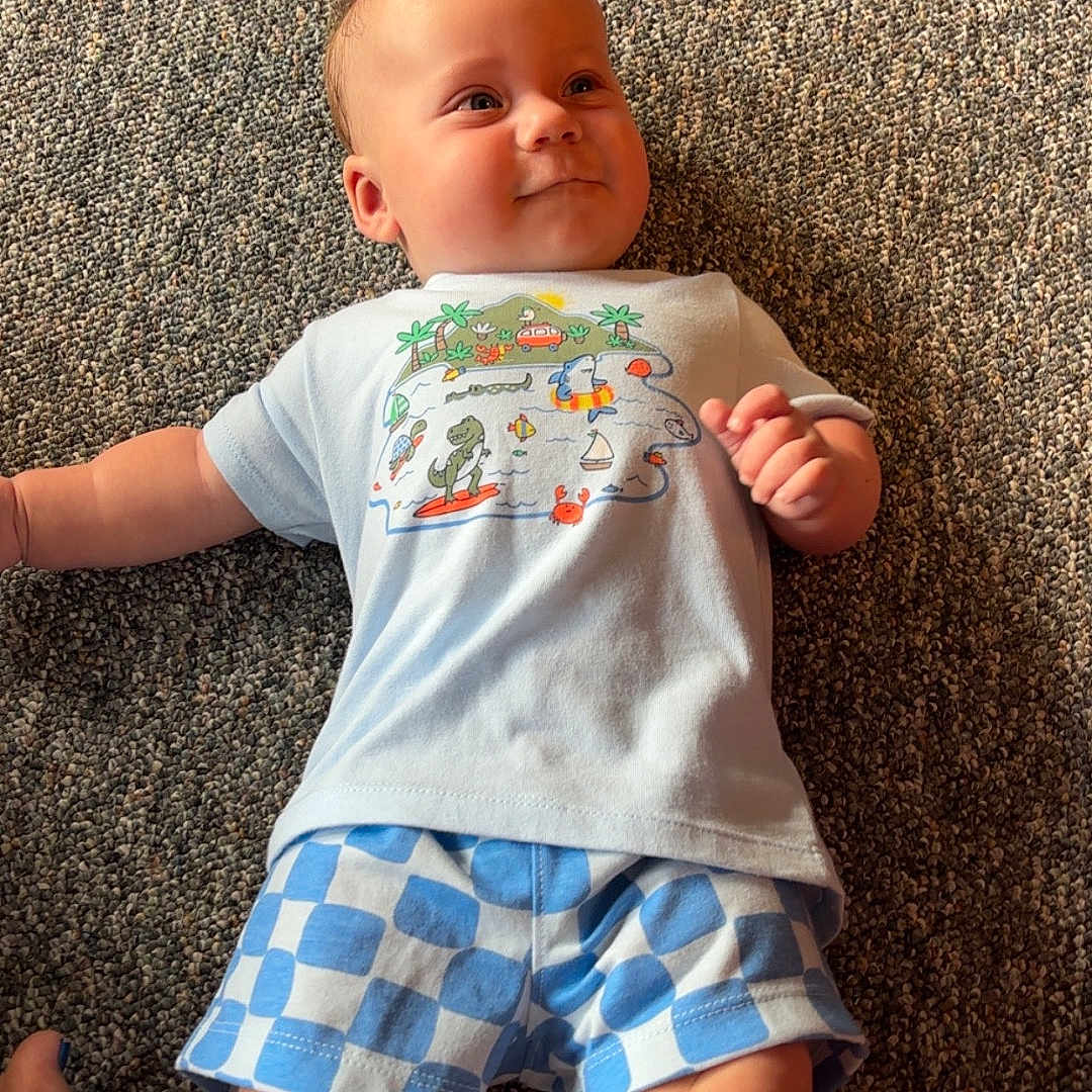 Garrett is registered to the contest to win money with this photo: baby, blue, carpet, cartoon, child, clothing, cute, floor, happy, indoor, infant, legs, lying_down, pattern, person, shorts, skin, smile, tshirt, white