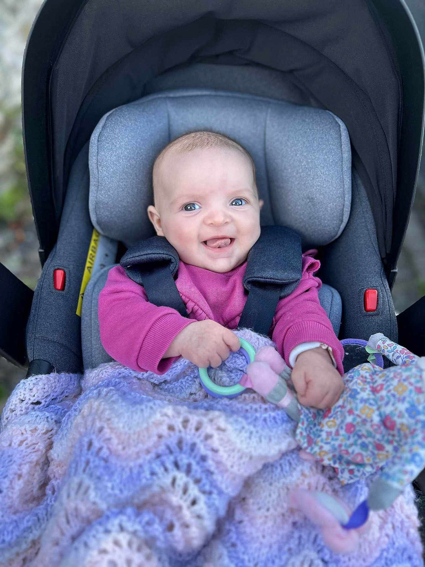 Sienna is registered to the contest to win money with this photo: baby, car_seat, blanket, toy, smile, happy, child, infant, face, clothing, purple, pink, knitted, portrait, cute, young_child, seated, hands, blue_eyes, indoor
