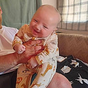 Maximus is registered to the contest to win money with this photo: baby, child, adult_hand, pajamas, animal_print, smile, blur, indoor, living_room, curtain, blanket, furniture, person, hand, leg, cozy, pattern, cute, happy, holding