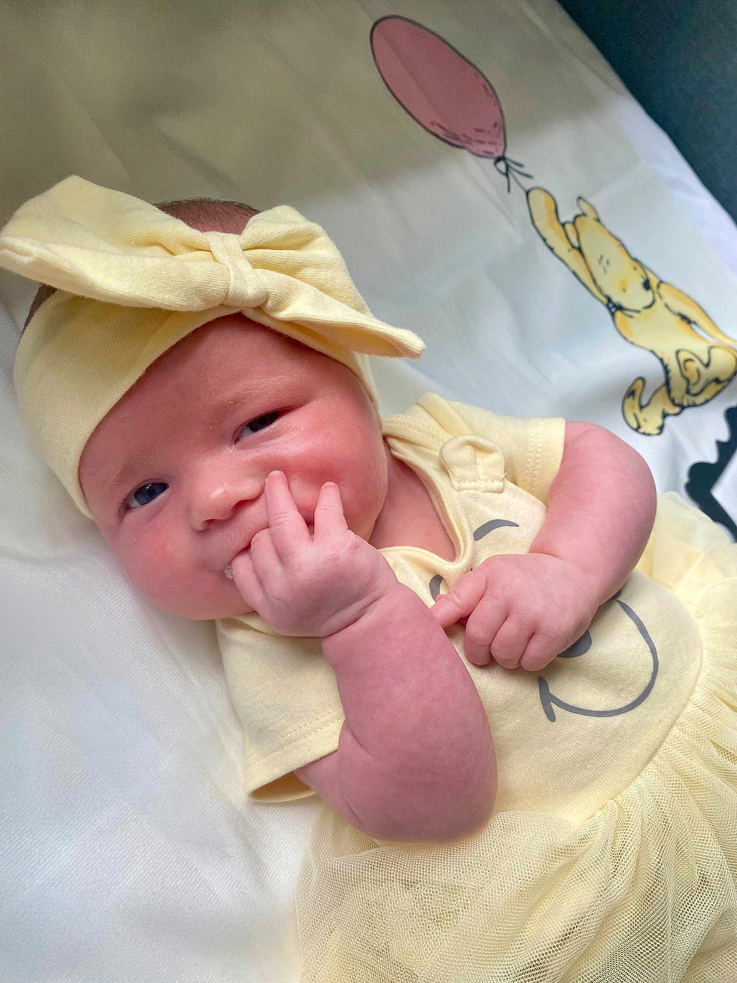 Rylee is registered to the contest to win money with this photo: baby, infant, child, yellow_clothing, headband, bow, hand, face, smiling_face, lying_down, blanket, cartoon, balloon, cute, skin, toddler, indoors, soft_texture, happy, portrait