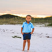 Levi joined the competition — help win amazing prizes! child, boy, beach, sand, sunset, smiling, barefoot, shorts, polo_shirt, blue_clothing, nature, outdoor, grass, dunes, sky, clouds, portrait, standing, casual, summer