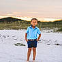 child, boy, beach, sand, sunset, smiling, barefoot, shorts, polo_shirt, blue_clothing, nature, outdoor, grass, dunes, sky, clouds, portrait, standing, casual, summer