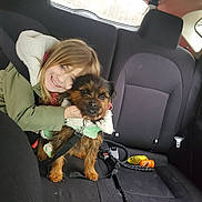 Bear is registered to the contest to win money with this photo: child, dog, car_interior, backseat, seatbelt, smiling, pet, leash, toy, jacket, young_girl, animal, happy, window, seat, black_seat, blond_hair, hug, clothing, domestic_animal