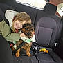 child, dog, car_interior, backseat, seatbelt, smiling, pet, leash, toy, jacket, young_girl, animal, happy, window, seat, black_seat, blond_hair, hug, clothing, domestic_animal