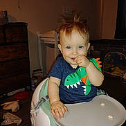 Ransom is registered to the contest to win money with this photo: baby, child, high_chair, dinosaur_shirt, blue_eyes, snack, indoor, furniture, wooden_floor, toy, cabinet, clothing, person, cute, smiling, hair, face, room, childhood, playpen