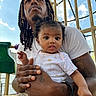 Wynterr is registered to the contest to win money with this photo: adult, baby, child, person, playground, outdoor, sky, clouds, clothing, tattoo, hair, hand, holding, expression, daylight, fence, metal, rope, white_shirt, curly_hair