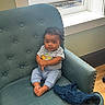 Ezekiel is registered to the contest to win money with this photo: armchair, baby, blanket, blue_clothing, child, cozy, curly_hair, cute, dinosaur_print, furniture, home, indoor, infant, person, portrait, relaxed, seated, smiling, sunlight, window