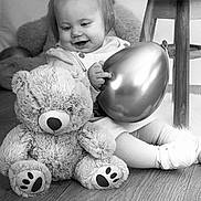 Lyna participe au concours pour gagner de l'argent avec cette photo : toddler, child, balloon, teddy_bear, stuffed_animal, pigtails, smiling, black_and_white, wooden_floor, table, furniture, indoor, happy, cute, toy, baby_shoes, seated, portrait, playful, infant
