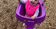 Lyra joined the competition — help win amazing prizes! baby, child, playground, swing, smiling, happy, outdoor, portrait, face, infant, pink_clothing, purple_swing, swing_chains, wood_chips, playground_equipment, sitting, gripping, daylight, adult_legs, recreation