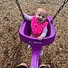 baby, child, playground, swing, smiling, happy, outdoor, portrait, face, infant, pink_clothing, purple_swing, swing_chains, wood_chips, playground_equipment, sitting, gripping, daylight, adult_legs, recreation