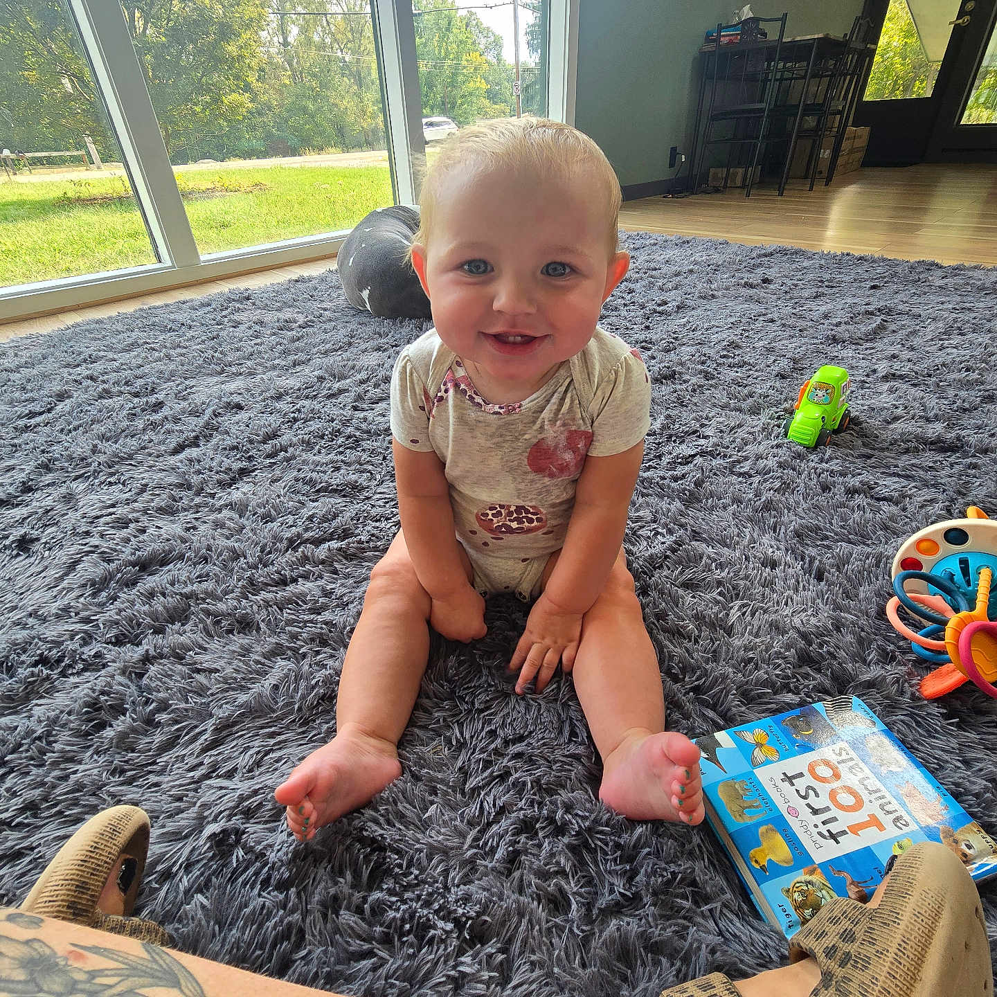 Lyra is registered to the contest to win money with this photo: baby, child, toddler, smiling, sitting, indoors, living_room, rug, toy_truck, activity_toy, picture_book, window, sunlight, playtime, onesie, barefoot, portrait, happy, home, tattoo