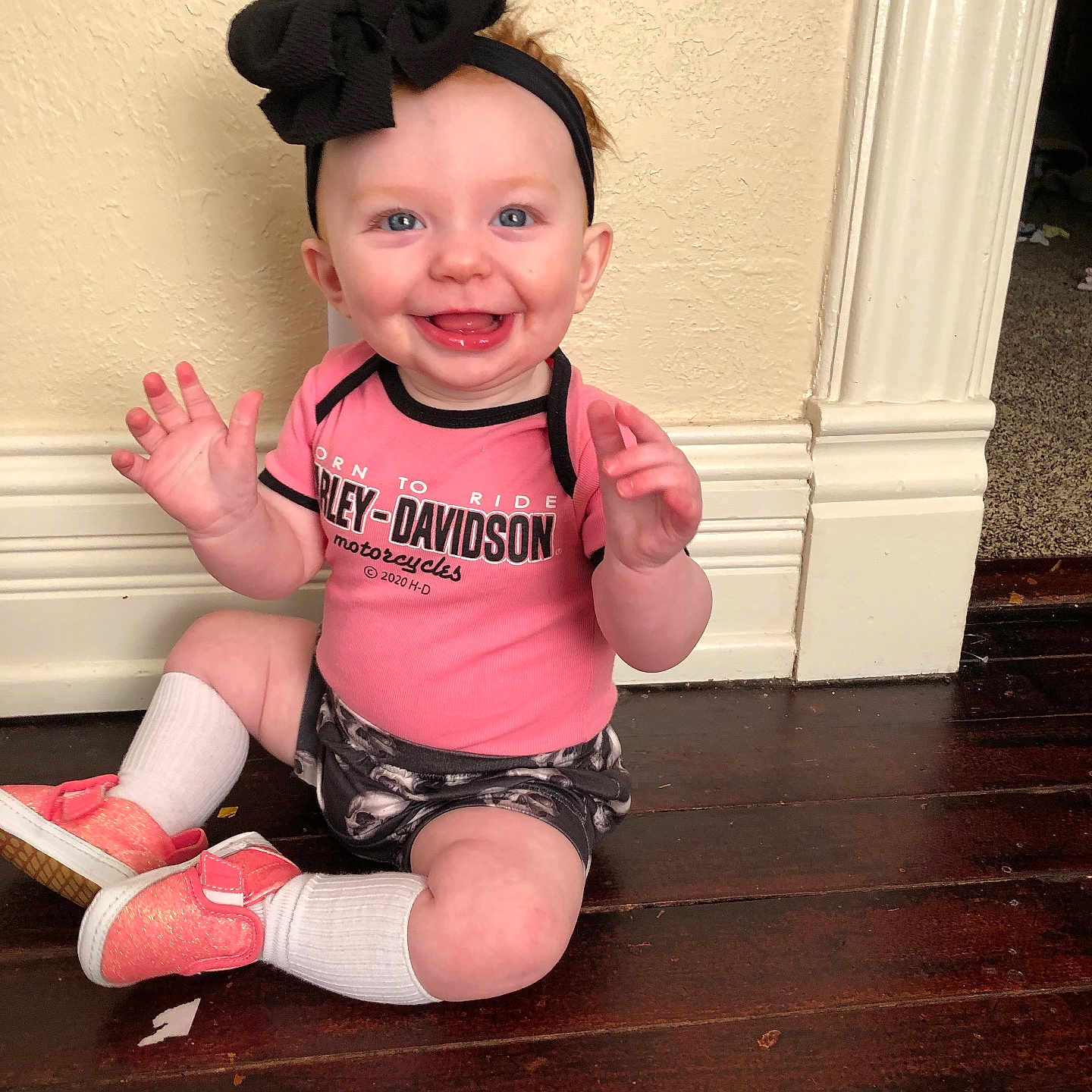 Ember joined the competition — help win amazing prizes! baby, baseboard, bow, child, cute, floor, hands, happy, harley_davidson, headband, indoors, person, pink_shirt, portrait, shoes, sitting, smiling, wall, white_socks, wooden_floor