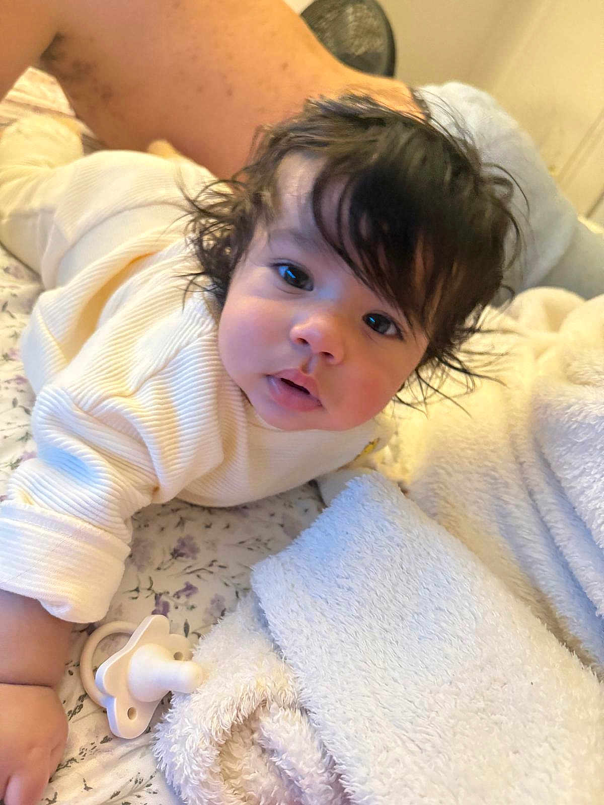 Javier joined the competition — help win amazing prizes! baby, infant, face, eyes, hair, pacifier, blanket, bedspread, clothing, soft, curly_hair, skin, indoors, person, child, lying_down, cute, young, resting, comfort