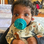 Mecca is registered to the contest to win money with this photo: baby, pacifier, curly_hair, onesie, animal_print, hand, indoor, wide_eyes, infant, child, cute, face, clothing, person, portrait, holding, expression, comfort, young, cozy