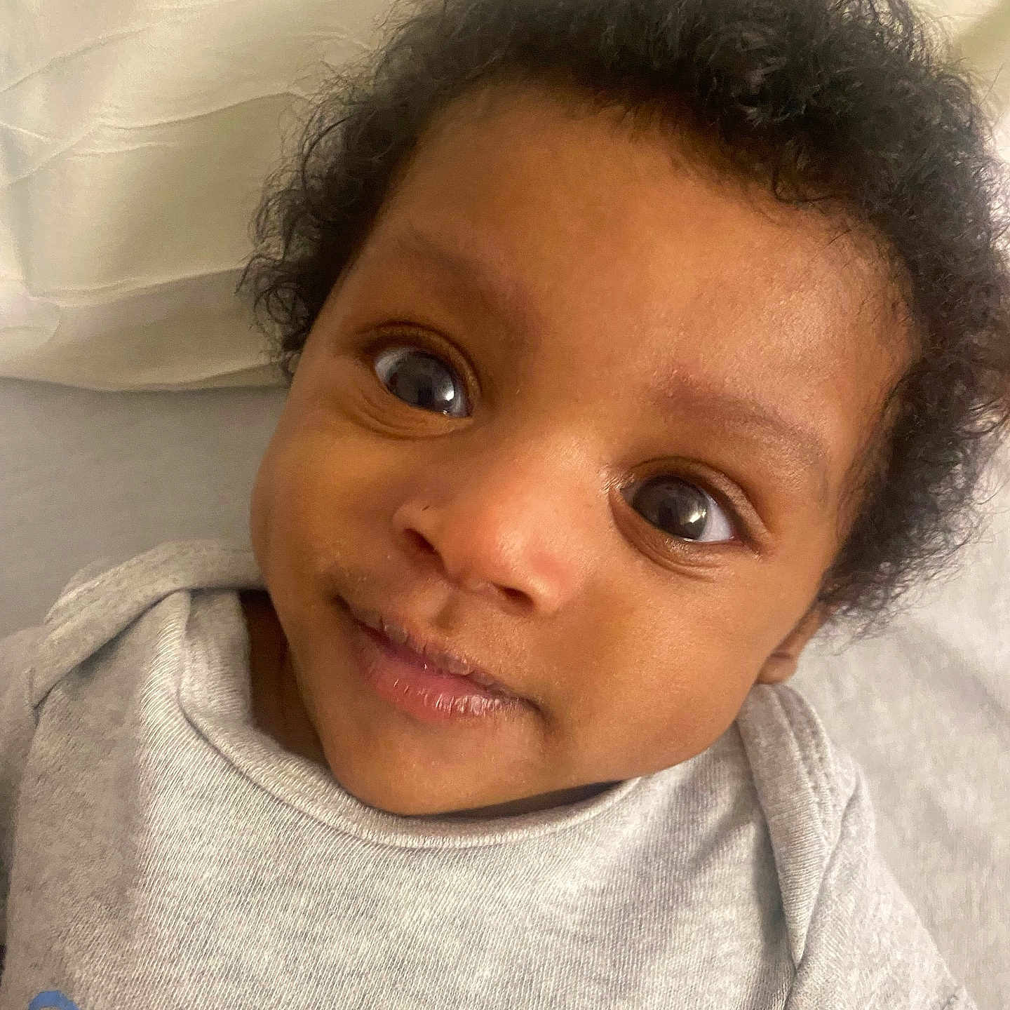 Mecca is registered to the contest to win money with this photo: baby, child, closeup, clothing, curly_hair, cute, expression, eyes, face, indoors, infant, lying_down, onesie, person, pillow, portrait, skin, smile, soft_surface, young_child