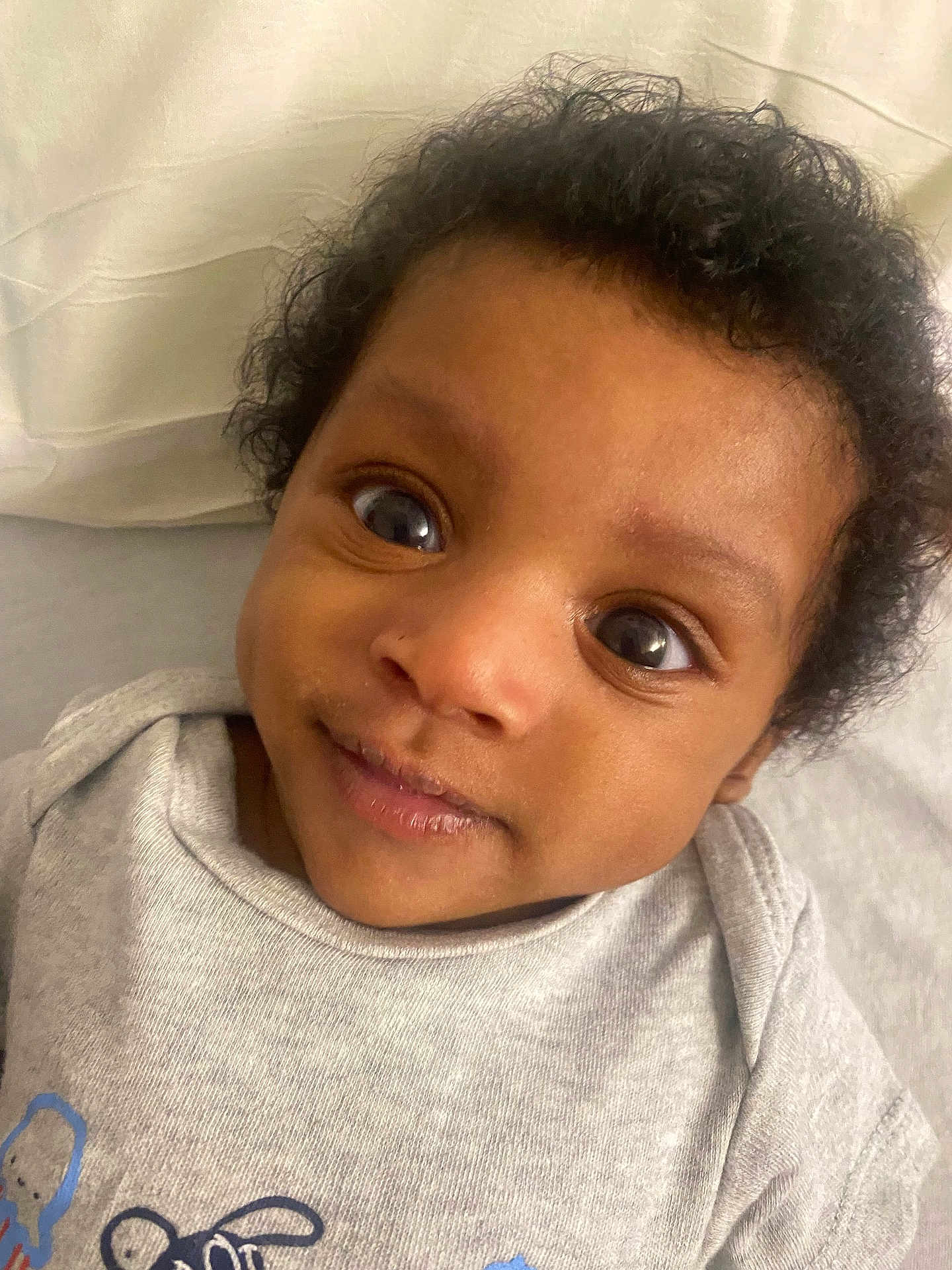 Mecca is registered to the contest to win money with this photo: baby, infant, face, eyes, smile, onesie, clothing, curly_hair, portrait, closeup, child, cute, lying_down, soft_surface, pillow, skin, young_child, indoors, expression, person